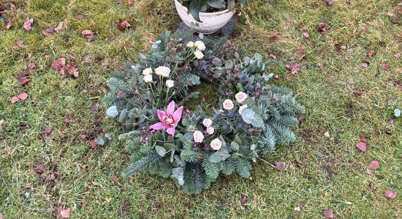 Christmas grave wreath of seasonal foliage and white and pink flowers laid in front of a gravestone and potted plant.

