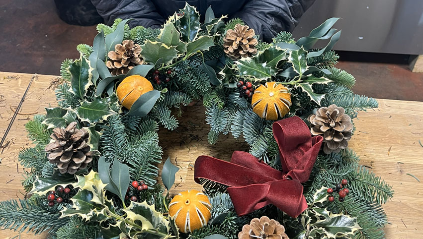 Christmas door wreath complete with holly, berries, pine cones, dried oranges, and a red velvet bow on a wooden work surface.