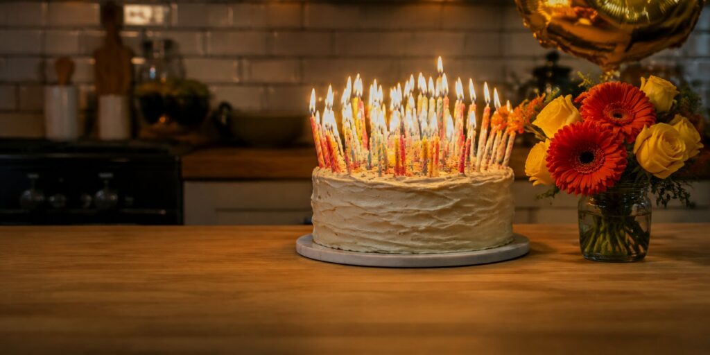 A birthday cake crowded with lit candles beside a small posy of orange and yellow flowers